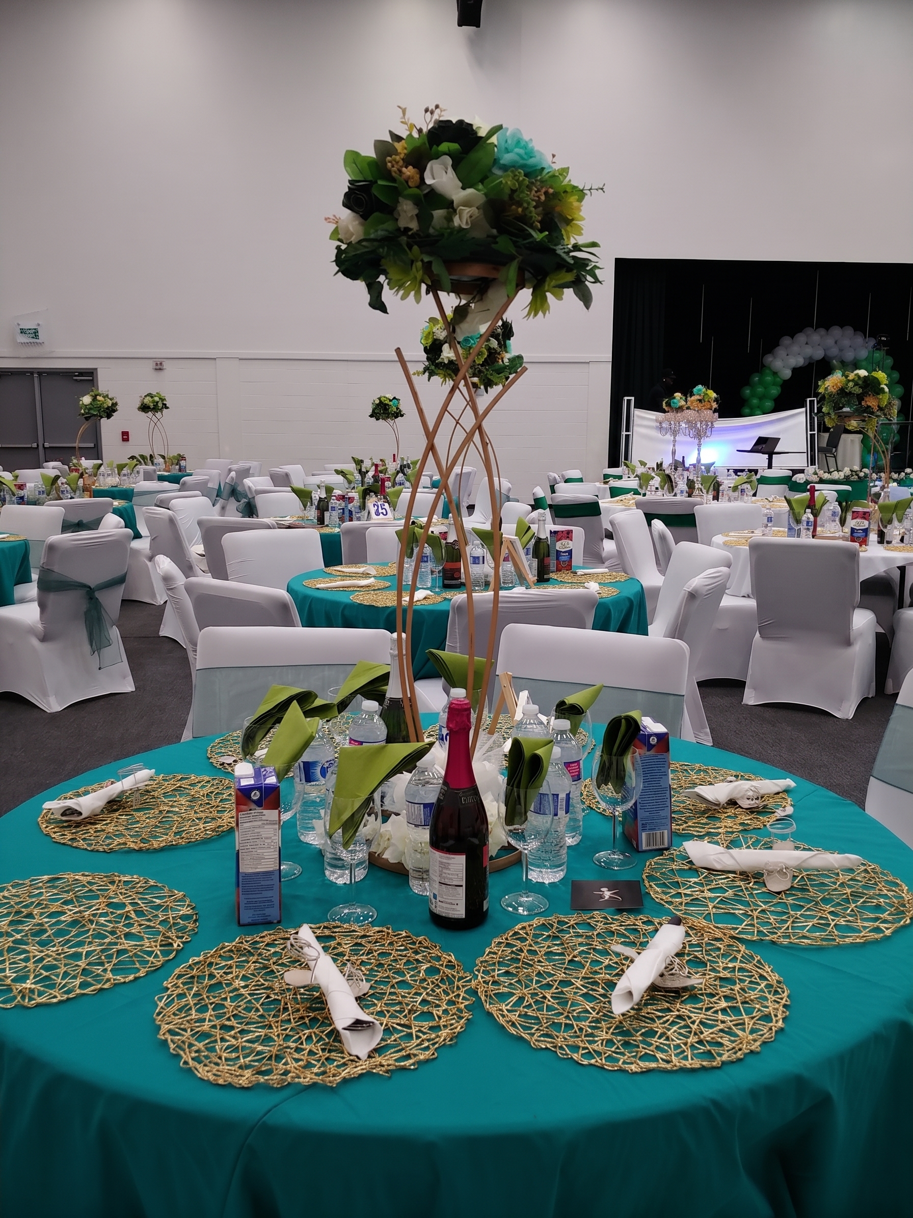 Teal and gold reception hall with tall floral centerpieces and a balloon arch on stage